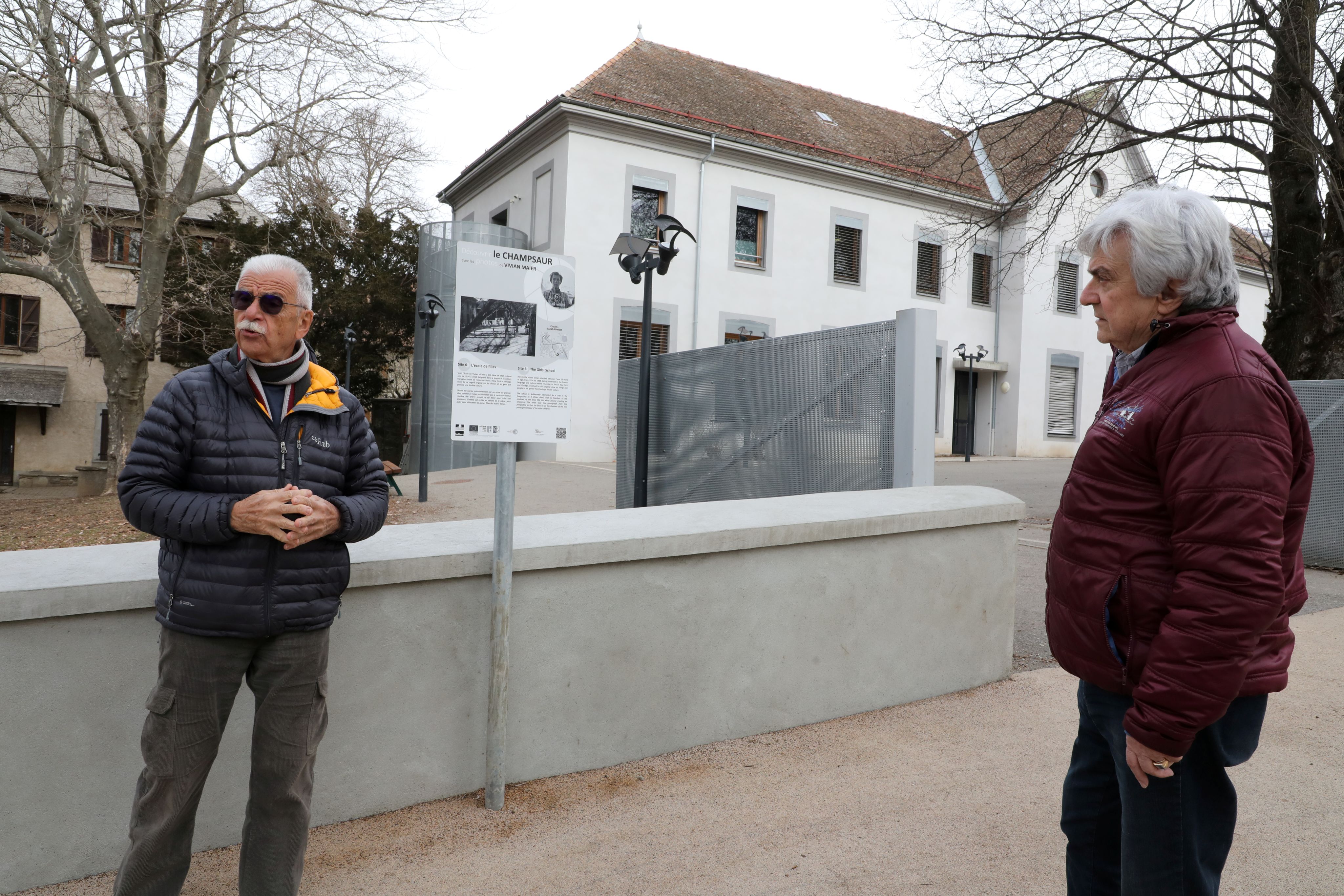 1. Alain Robert, trésorier de l’association et photographe amateur, en compagnie de Tony Gay, secrétaire. Les deux hommes sont devant l’un des panneaux installés pour raconter l’histoire des images de Vivian Maier. Celui-ci est posé devant la Maison Blanche, bâtiment qui abritait autrefois l’école dans laquelle l’artiste était scolarisée en 1932.